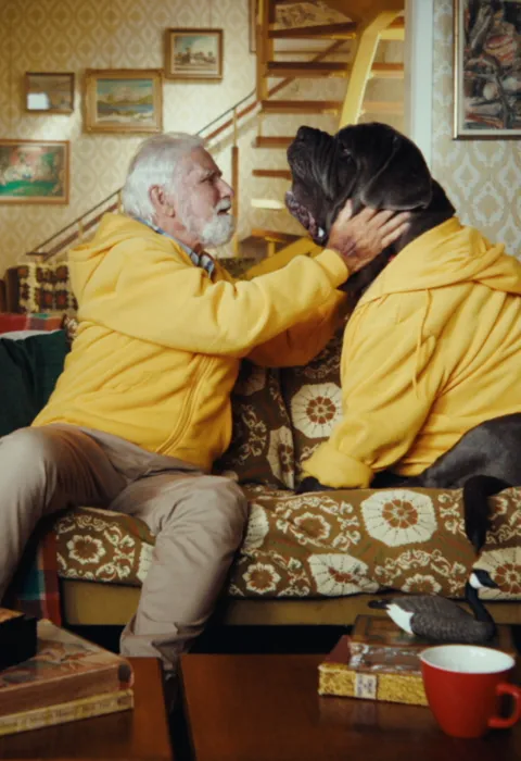 Man with white hair and wearing a yellow jacket, hugging a dog also in a yellow jacket.