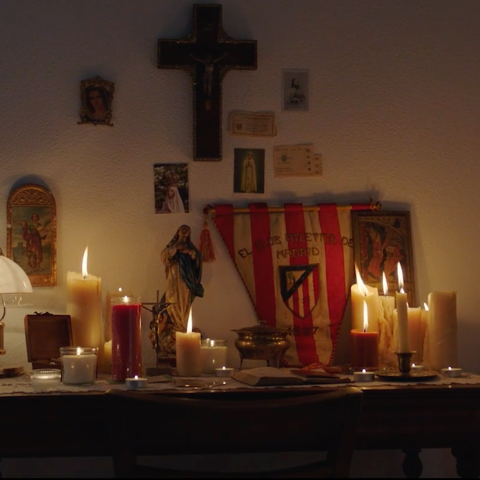 A home altar filled with lit candles, religious icons, and an Atlético de Madrid flag displayed beneath a crucifix.
