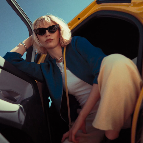 Woman wearing sunglasses sits in the open door of the Ford Capri EV, photographed from a low angle against a blue sky.