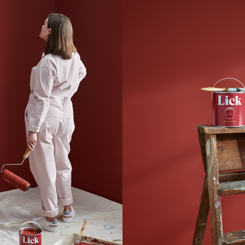 Woman in light overalls standing on protective sheets, holding a paint roller in front of a red wall with Lick x Heinz paint tin on the floor.