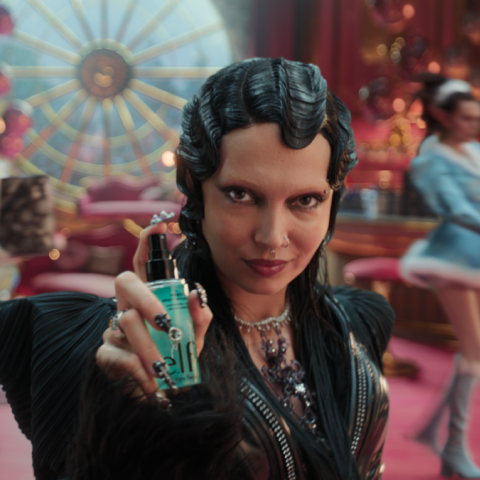 Woman in a dramatic black outfit presenting a beauty serum, with dancers and Christmas décor in a stylised, glamorous holiday scene.