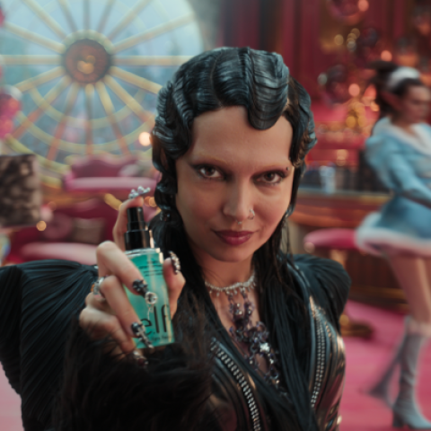 Woman with sculpted vintage hairstyle holding a teal beauty serum bottle in a festive, theatrical Christmas setting.