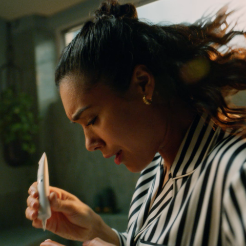Woman in striped pyjamas examining and applying skincare serum in softly lit bathroom.