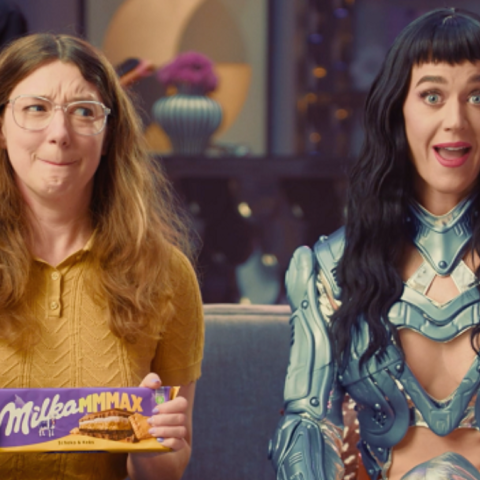 Woman holding Milka MMMAX chocolate bar sitting beside popstar styled character wearing futuristic metallic armour on a sofa.