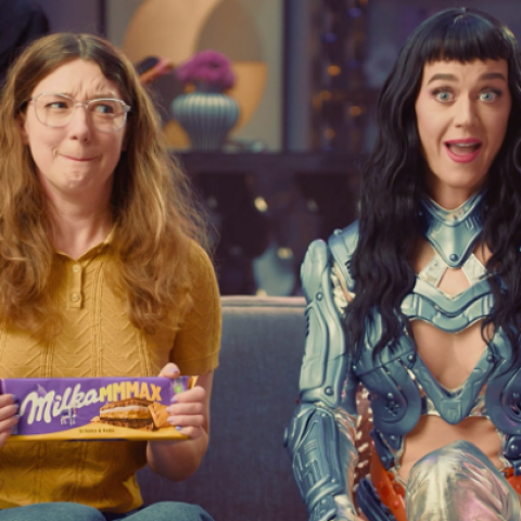 Woman holding Milka MMMAX chocolate bar sitting next to futuristic popstar style character reacting with surprise.