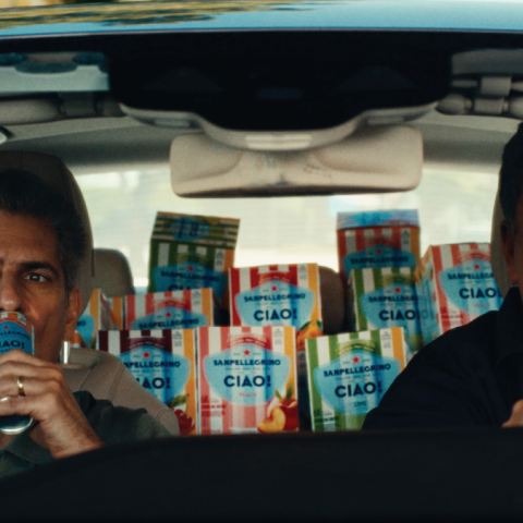 Two men sitting in a car filled with Sanpellegrino CIAO! multipacks, one drinking a can while the other drives, with colourful CIAO! packaging visible in the back seat.